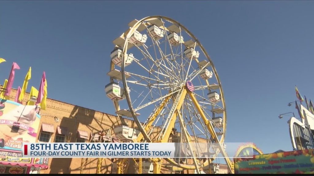 A large yellow Ferris wheel at the 85th East Texas Yamboree four-day county fair in Gilmer, Texas, set against a clear blue sky with fair food booths in the background.