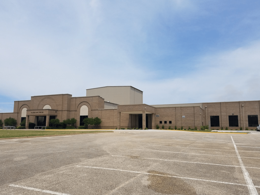 The exterior of the Gilmer Civic Center, a large tan brick multi-purpose building with a spacious parking lot in Gilmer, Texas.
