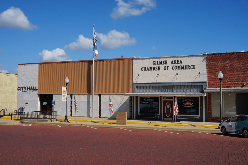 A view of the Gilmer City Hall and Gilmer Area Chamber of Commerce buildings on a brick-paved street in Gilmer, Texas, featuring American flags and a clear blue sky.