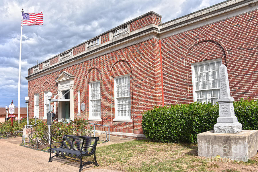 The exterior of the Historic Upshur Museum, a red brick building in Gilmer, Texas, featuring arched windows, an American flag on a tall flagpole, and a stone memorial monument in the foreground.