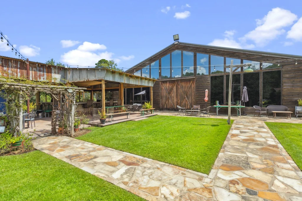 The outdoor patio and tasting area at Los Pinos Ranch Vineyards in Gilmer, Texas, featuring a rustic wooden barn with large glass windows, stone walkways, and a lush green lawn under a blue sky.