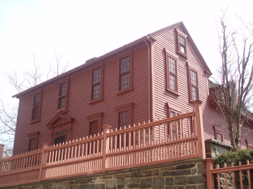 he exterior of the historic Paul Revere House, a three-story colonial wooden building with a red facade and a matching picket fence, situated on a stone foundation in Boston, Massachusetts.
