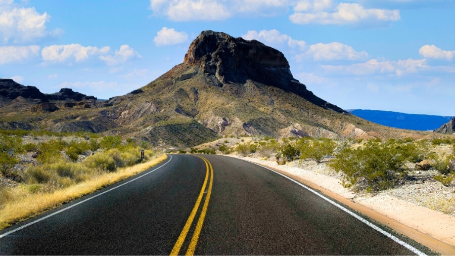 A paved highway with double yellow lines winding through a desert landscape in West Texas, leading toward a large, rocky mountain peak under a bright blue sky with scattered clouds.