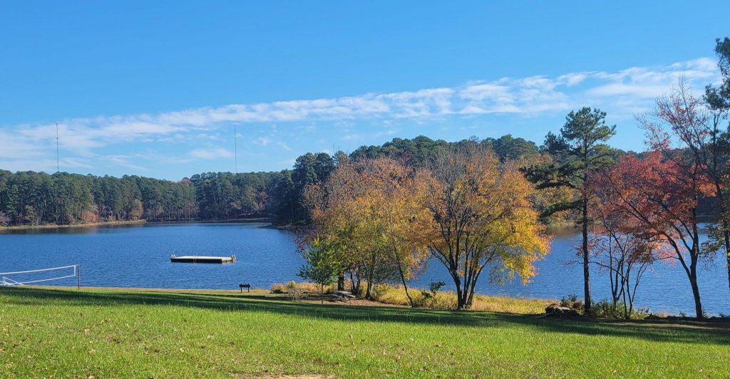 A tranquil view of the 80-acre Little Pine Lake at Daingerfield State Park, featuring a small wooden pier and a white boat on the calm water, surrounded by the dense pine and hardwood forests of East Texas near Gilmer.