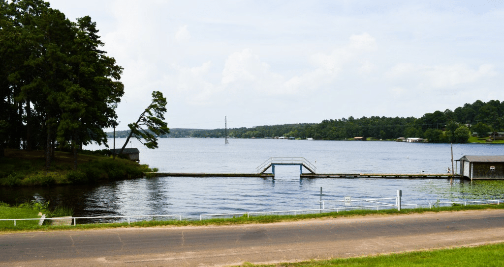 A wide view of the serene Lake Gladewater near Gilmer, Texas, featuring a calm waterfront with a small walking bridge, surrounded by lush green trees and local boat houses under a bright afternoon sky.