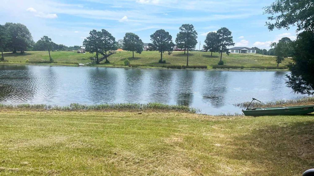 A scenic view of a calm lake in Longview, Texas, featuring a grassy shoreline with a small green boat, a wooden staircase leading up a grassy hill with pine trees, and a clear blue sky, highlighting a peaceful outdoor destination near Gilmer.