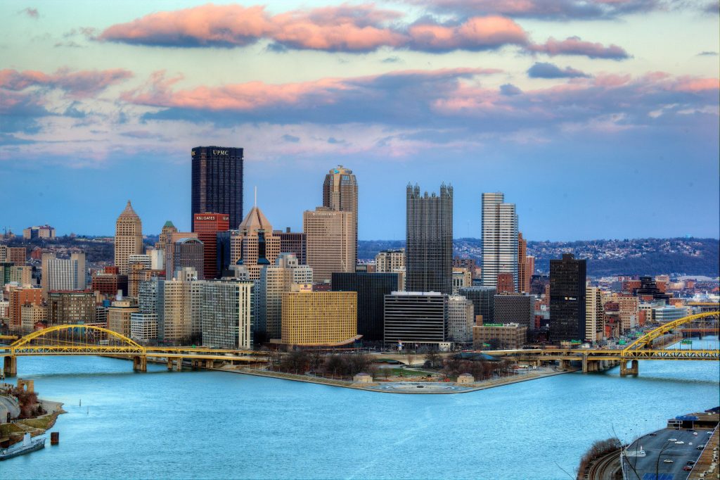 A wide panoramic view of the Pittsburgh, Texas skyline featuring modern high-rise buildings situated at the confluence of two rivers with yellow bridges, illustrating a unique urban day trip destination near Gilmer.